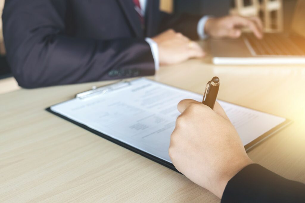 Close up of a hand filling out paperwork with another person working on a computer sitting across from them.